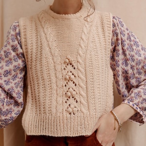 以下が含まれることがあります： ケーブル編みのパターンと繊細な透かし模様が施されたクリーム色のニットベスト。ベストは丸首で、丈が短くなっています。