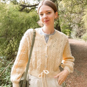 May include: A pale yellow knitted cardigan with a textured design and decorative buttons. The cardigan has long sleeves and a drawstring waist. The person is wearing light-coloured trousers and a green shoulder bag.