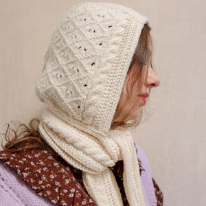 Op de afbeelding: Een witte gebreide capuchon sjaal met een kabelbreipatroon. De sjaal wordt gedragen door een persoon met lang bruin haar.