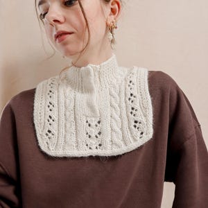 May include: A white knitted cowl neck with a cable knit pattern and delicate lace details. The cowl is worn over a brown turtleneck sweater.
