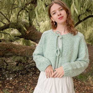 May include: A pale green knitted cardigan with a tie fastening, worn with a white tiered skirt. The sweater has long sleeves and a round neck. The background shows a large tree with green leaves.
