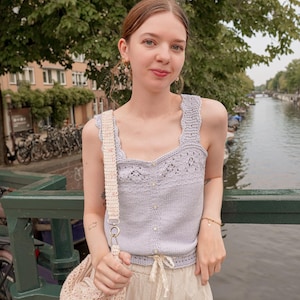 Op de afbeelding: Een lichtpaarse gehaakte tanktop met een geschulpte halslijn en knoopsluiting, gedragen met een beige broek. Een bijpassende gehaakte tas met een crèmekleurige band is ook zichtbaar. De achtergrond toont een gracht en bomen.