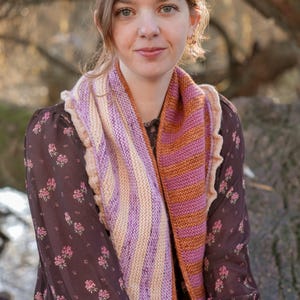 Op de afbeelding: Een vrouw draagt een kleurrijke gebreide sjaal met verticale strepen in tinten paars, oranje en crème. De sjaal is om de nek en borst gedrapeerd. Ze draagt een donkerbruine jurk met bloemenprint. De achtergrond is wazig met bomen.