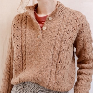 Op de afbeelding: Een bruine gebreide trui met een kabelbreiselpatroon en een enkele knoopsluiting bij de hals. De trui heeft een ontspannen pasvorm en is gemaakt van een zacht, pluche materiaal.