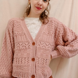 Puede incluir: Un cárdigan de crochet rosa con cuello en V y cierre de botones. El cárdigan tiene un delicado patrón de calado y una longitud ligeramente recortada.