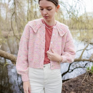 Op de afbeelding: Een roze en witte cardigan met een grote kraag en parelknopen. De cardigan is gemaakt van een zacht, pluche materiaal en heeft een licht getailleerde pasvorm. Het model draagt de cardigan over een rood T-shirt en een witte jeans.