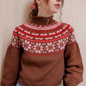 May include: A brown knitted sweater with a pink and red geometric pattern. The sweater has a high neck and long sleeves.