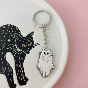 May include: A white ghost keychain with a black cat illustration on a white plate. The ghost has a cute face with two eyes and a small mouth. The keychain is attached to a silver key ring.