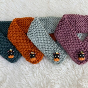 May include: Four knitted neck warmers in different colors: teal, orange, gray, and purple. Each warmer has a black cat with a pumpkin on its head.