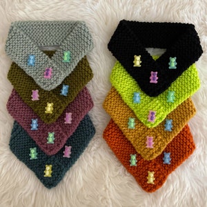 Peut inclure: Une pile de dix chauffe-cou en tricot de différentes couleurs, dont le gris, le noir, le vert, le marron, le marron et l'orange. Chaque chauffe-cou est orné d'un charme en forme d'ours en gélatine.