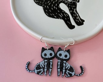 May include: A pair of black cat skeleton earrings with white bone details. The earrings are hanging from silver hooks.