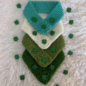 May include: Four knitted neck warmers in shades of green and white, with shamrock embellishments. The neck warmers are stacked on top of each other, with the green ones on the bottom and the white one on top.