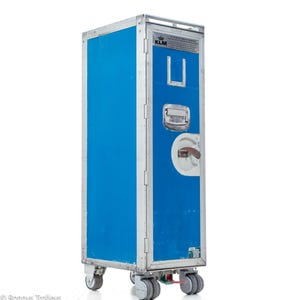 Peut inclure: Chariot de service de la compagnie aérienne KLM bleu et argenté, à quatre roues. Le chariot a une porte bleue avec un cadre argenté et une poignée. Le logo KLM est visible en haut.