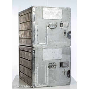 May include: Two stacked, industrial-style metal storage containers. The containers are silver with riveted edges, horizontal ribbed detailing, and a handle. Each has a rectangular opening and the word "Britannia" is visible on the front.