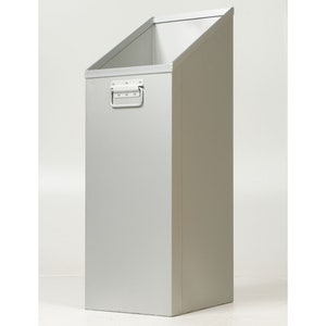 May include: A tall, silver-colored metal wastebasket with a slanted top and a handle. The rectangular bin has a handle on the front and is designed to stand upright. The interior is visible, and the overall design is minimalist and functional.