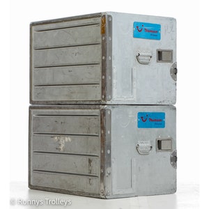 May include: Two stacked, rectangular, silver-colored metal storage containers. Each container has a handle, a small rectangular opening, and a blue sticker with the text "Thomson Airways". The containers appear to be used and have some wear and tear.