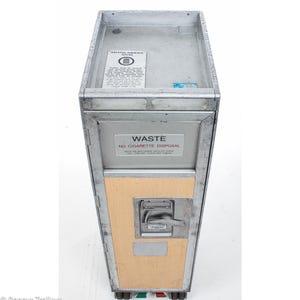 May include: A vintage British Airways Atlas aircraft trolley. The metal frame surrounds a wooden panel and features a "Waste - No Cigarette Disposal" sign. The top has a British Airways sticker. The trolley has wheels with red and green accents.