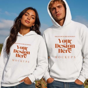 May include: Two white hooded sweatshirts featuring the text Millennium Dream Design Your Design Here Mockups in orange lettering. The hoodies have long sleeves, a front kangaroo pocket, and a drawstring hood, worn over blue denim jeans.