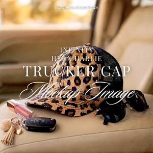 May include: A leopard print trucker cap with a black mesh back, accompanied by black sunglasses, a pink lip gloss tube, and a keychain with a small beige pom-pom and a tassel. The text INFINITY HER CHARLIE TRUCKER CAP Mockup Image is visible.