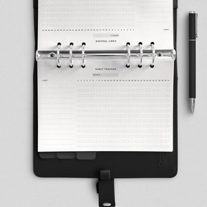 May include: A black binder with a white habit tracker page inside. The page has a grid of squares for tracking daily habits. The text "HABIT TRACKER" is printed at the top of the page.