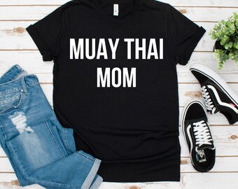 May include: Black t-shirt with the text "MUAY THAI MOM" in white block letters. The shirt is displayed with a pair of blue jeans and black and white sneakers. A small green plant is also visible.