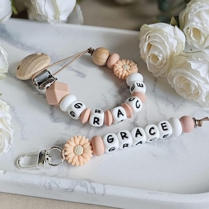 May include: A personalised baby pacifier clip and matching keychain. The pacifier clip and keychain are white, peach, and wooden beads. The keychain has the name "GRACE" spelled out in white letter beads. The pacifier clip has the name "GRACE" spelled out in white letter beads.