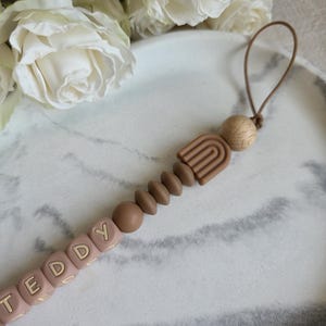 May include: A baby pacifier clip with the name "TEDDY" spelled out in light pink letter beads. The clip features a brown rainbow design, wooden beads, and a loop for attachment. The background includes white roses and a marble-patterned surface.