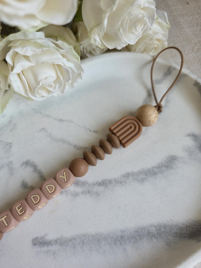 May include: A baby pacifier clip with the name "TEDDY" spelled out in light pink letter beads. The clip features a brown rainbow design, wooden beads, and a loop for attachment. The background includes white roses and a marble-patterned surface.