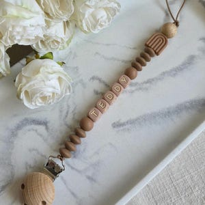 May include: A baby pacifier clip with the name "TEDDY" spelled out in light brown letter beads. The clip features a wooden clip, brown beads, and a small rainbow design. The pacifier clip is lying on a white tray with white roses in the background.