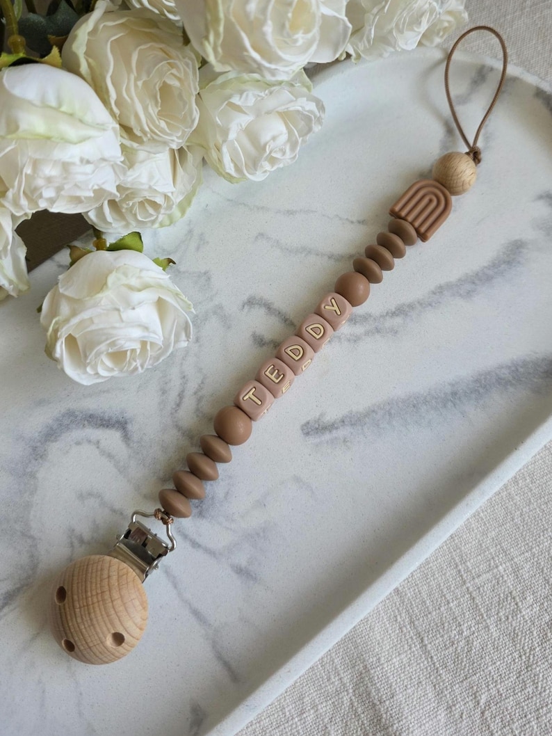 May include: A baby pacifier clip with the name "TEDDY" spelled out in light brown letter beads. The clip features a wooden clip, brown beads, and a small rainbow design. The pacifier clip is lying on a white tray with white roses in the background.