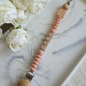 May include: A baby pacifier clip with the name "ZARA" spelled out in wooden letter beads. The clip features a wooden rainbow, wooden beads, and a metal clip. The pacifier clip is pink and brown.