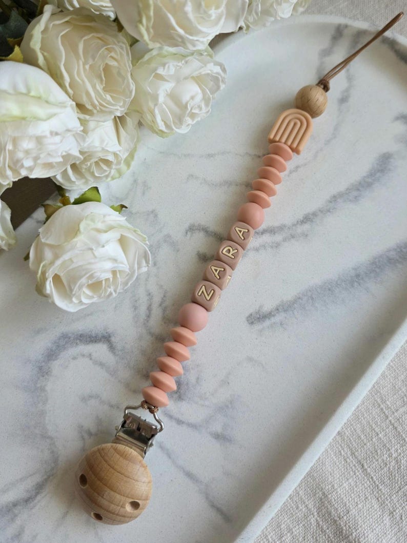 May include: A baby pacifier clip with the name "ZARA" spelled out in wooden letter beads. The clip features a wooden rainbow, wooden beads, and a metal clip. The pacifier clip is pink and brown.