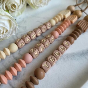 May include: Three baby pacifier clips in neutral colors, each with a different name spelled out in block letters: MARCUS, ZARA, and TEDDY. The clips feature a variety of bead shapes and sizes, including round and geometric designs. The background includes white roses.