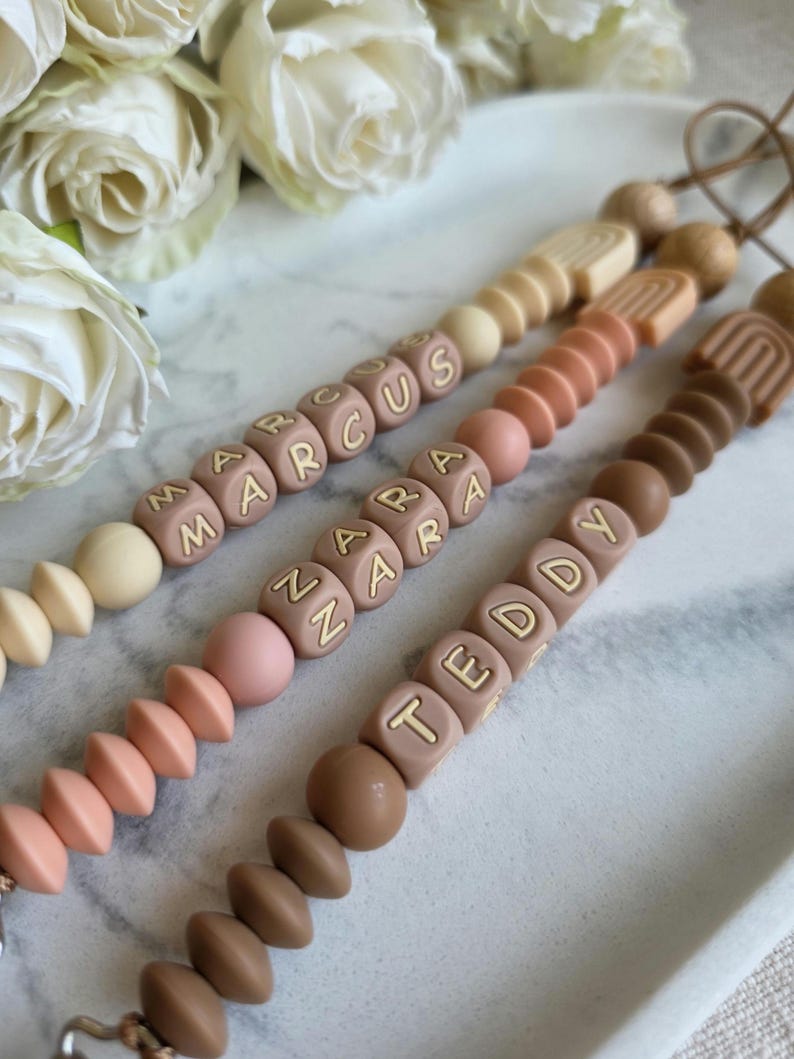 May include: Three baby pacifier clips in neutral colors, each with a different name spelled out in block letters: MARCUS, ZARA, and TEDDY. The clips feature a variety of bead shapes and sizes, including round and geometric designs. The background includes white roses.