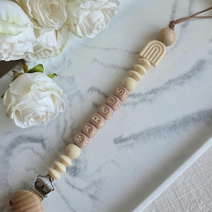 May include: A baby pacifier clip with the name "MARCUS" spelled out in pink letter beads. The clip features a wooden clip, wooden beads, and a beige rainbow design. The pacifier clip is attached to a brown cord.
