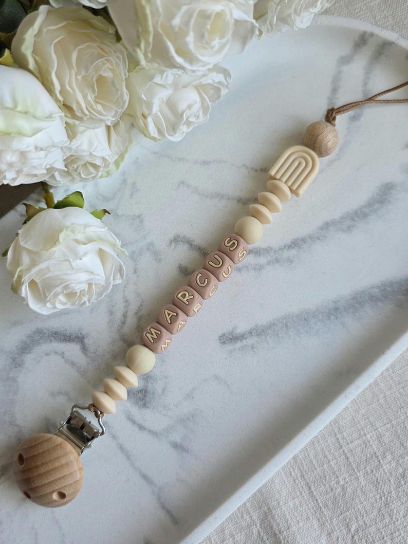 May include: A baby pacifier clip with the name "MARCUS" spelled out in pink letter beads. The clip features a wooden clip, wooden beads, and a beige rainbow design. The pacifier clip is attached to a brown cord.