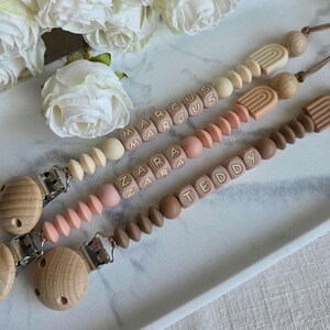 May include: Three personalized baby pacifier clips with wooden beads and the names "Marcus", "Zara", and "Teddy" spelled out in lettered blocks. The clips feature a variety of colors, including cream, pink, and brown, and are displayed on a white tray.