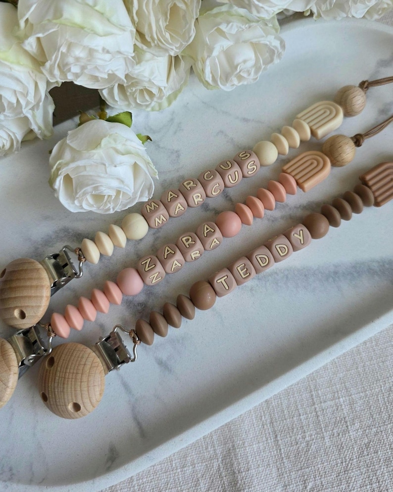 May include: Three personalized baby pacifier clips with wooden beads and the names "Marcus", "Zara", and "Teddy" spelled out in lettered blocks. The clips feature a variety of colors, including cream, pink, and brown, and are displayed on a white tray.