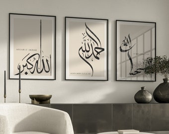 May include: Three framed prints with black calligraphy on a beige background. The prints feature the Arabic words "Allah-u-Akbar", "Alhamdulillah", and "Subhanallah". The prints are hanging on a white wall above a white armchair and a round coffee table.