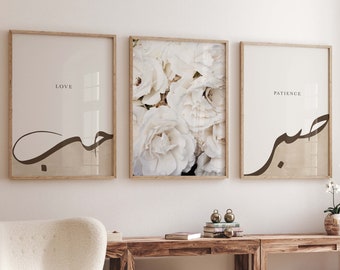May include: Three framed prints with white flowers in the center print and the words "LOVE" and "PATIENCE" in Arabic calligraphy on either side. The prints are hanging on a white wall above a wooden table.