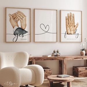 May include: Three framed prints with watercolor illustrations. The first print shows two hands holding each other with a ring on the woman's finger. The text "yes" is written below the hands. The second print shows a simple heart shape drawn with a black line. The third print shows four hands with the text "family" written below. The names "Rene & Tanja" and "Rene Tanja Maya Lya" are written below the text on the first and third prints.