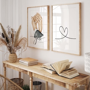 May include: Two framed prints, one with a watercolor illustration of two hands holding each other with a ring on the woman's finger and the word "yes" written below, and the other with a simple black line drawing of a heart. The date "15 November 2018" is written below the hands.