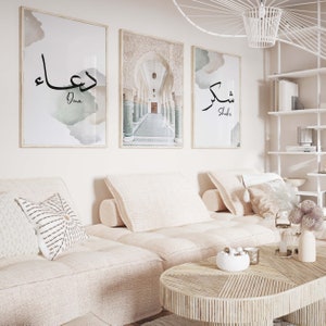 May include: Three framed prints with Arabic calligraphy and a photograph of an arched hallway. The prints are hung on a white wall above a light beige couch with white pillows.