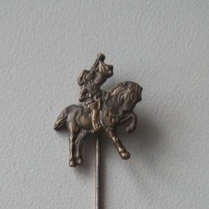 Könnte beinhalten: Vintage, silberfarbene Hutnadel mit detailliertem Reiterdesign. Die Nadel zeigt einen Reiter auf einem Pferd, mit aufwendigen Details an Pferd und Kleidung des Reiters. Die Nadel ist lang und schlank, mit einem strukturierten Schaft, etwa 10 cm lang.
