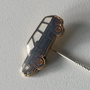 Può includere: Uno spillo dorato con un'immagine in bianco e nero di un'auto. Lo spillo ha un retro in metallo argentato.
