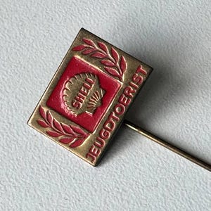 May include: A vintage pin badge with a red and gold design. The badge features the Shell logo and the text "JEUGDTOERIST" in gold lettering. The badge is diamond-shaped with a long, thin needle.