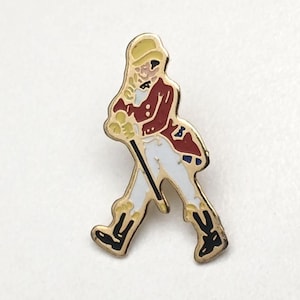 Op de afbeelding: Een goudkleurige emaille pin met een figuur in een rode jas, witte broek en zwarte laarzen, die een wandelstok vasthoudt. De figuur loopt, met een gele hoed. De pin staat op een witte achtergrond.
