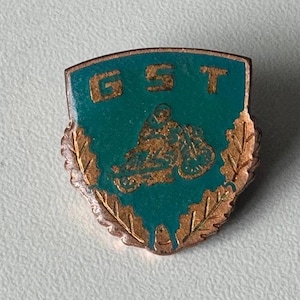 Op de afbeelding: Een schildvormige metalen speld met een groenblauwe emaille achtergrond en de letters GST in goud aan de bovenzijde. Een goudkleurige afbeelding van een persoon op een motorfiets staat centraal, omringd door een rand van eikenbladeren.