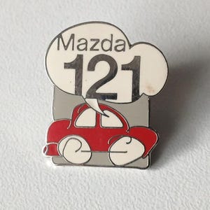 Könnte beinhalten: Ein Mazda 121 Auto-Pin. Der Pin zeigt ein rotes Auto-Design mit weißen Akzenten, einen grauen Hintergrund und eine Sprechblase mit dem Text "Mazda 121". Ein Sammlerstück.