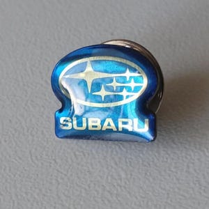 Könnte beinhalten: Ein blau-goldener Subaru-Emblem-Pin. Der Pin zeigt das Subaru-Logo mit blauem Hintergrund und goldenen Sternen. Das Wort "SUBARU" ist unten auf dem Pin in Weiß aufgedruckt.